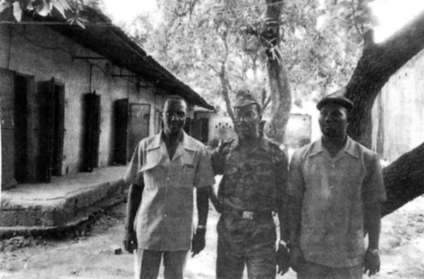 Bâtiment n°6. Avec, au fond, le réduit servant de local pour la vidange journalière. Alsény R. Gomez à gauche, pose ici en compagnie du Lieutenant-Colonel Kaba 41 Camara, au centre, qu'il omet de nommer par deux fois. Le troisième individu n'est pas identifié.